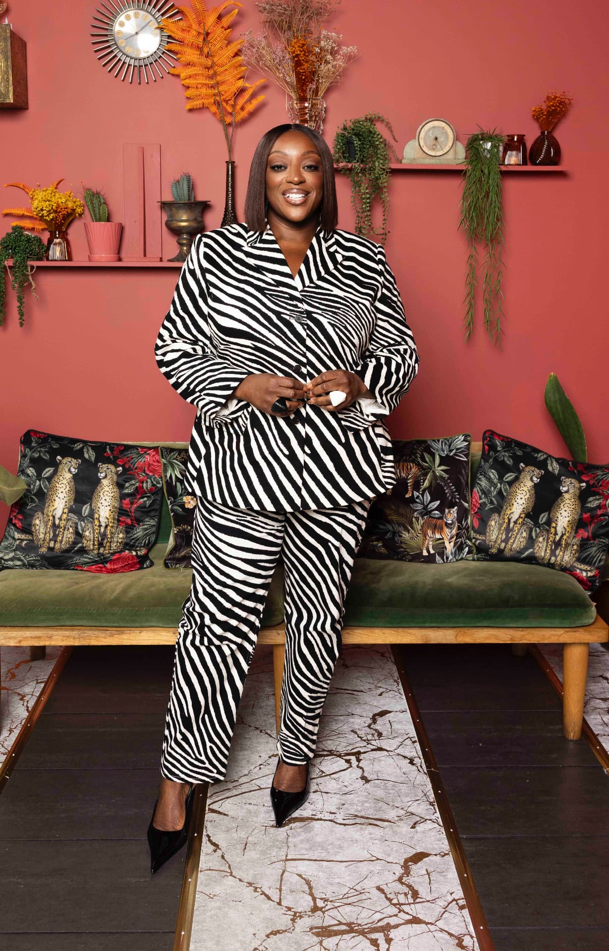 Judi Love smiling in a grey checked dress against a deep red wall